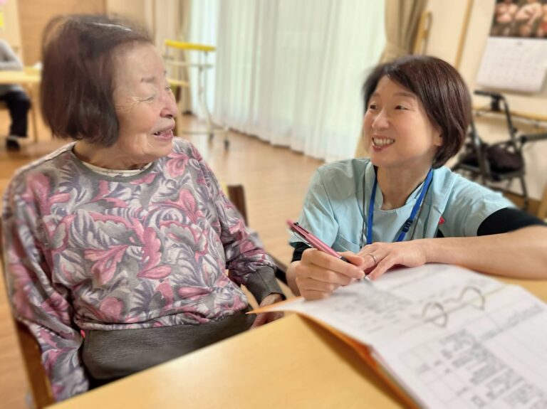 スタッフと入居者が机に向かい、書類を見ながら笑顔で会話している生活風景